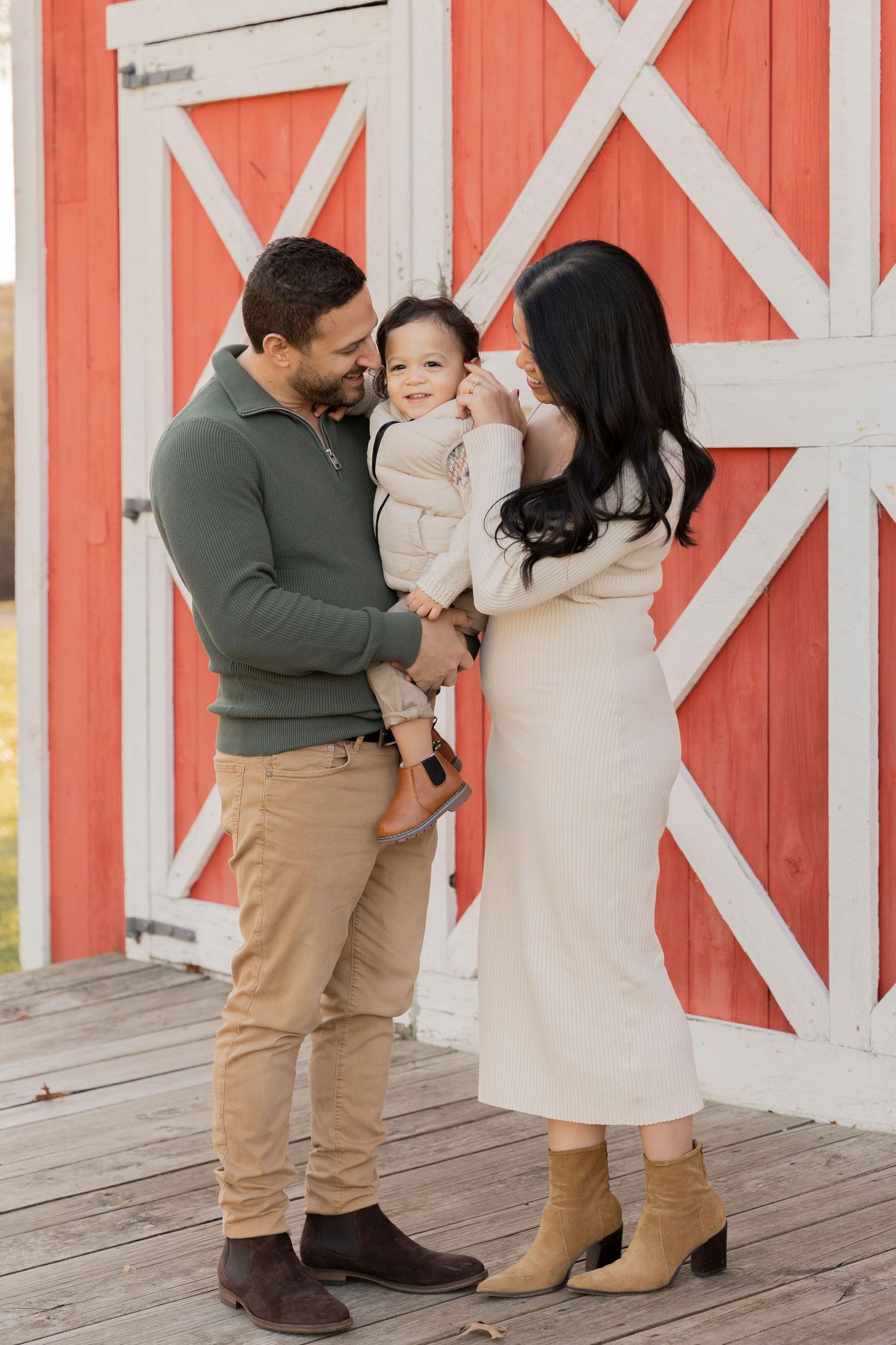 family of three smiling at their toddler boy in front of a red barn at cranberry lake park