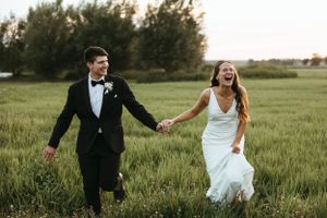 Joyful newlyweds running hand in hand through green field at sunset.
