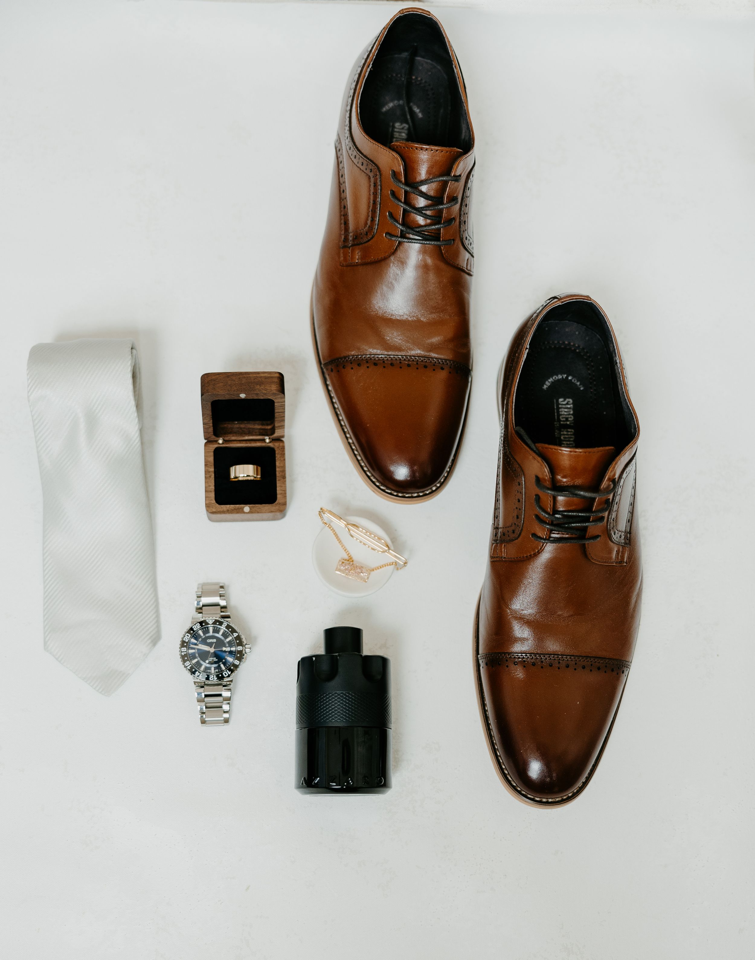 Groom's accessories flatlay with brown leather shoes, watch, tie, cologne and ring box.