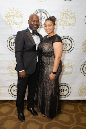 Couple in formal attire poses at elegant event with branded backdrop.