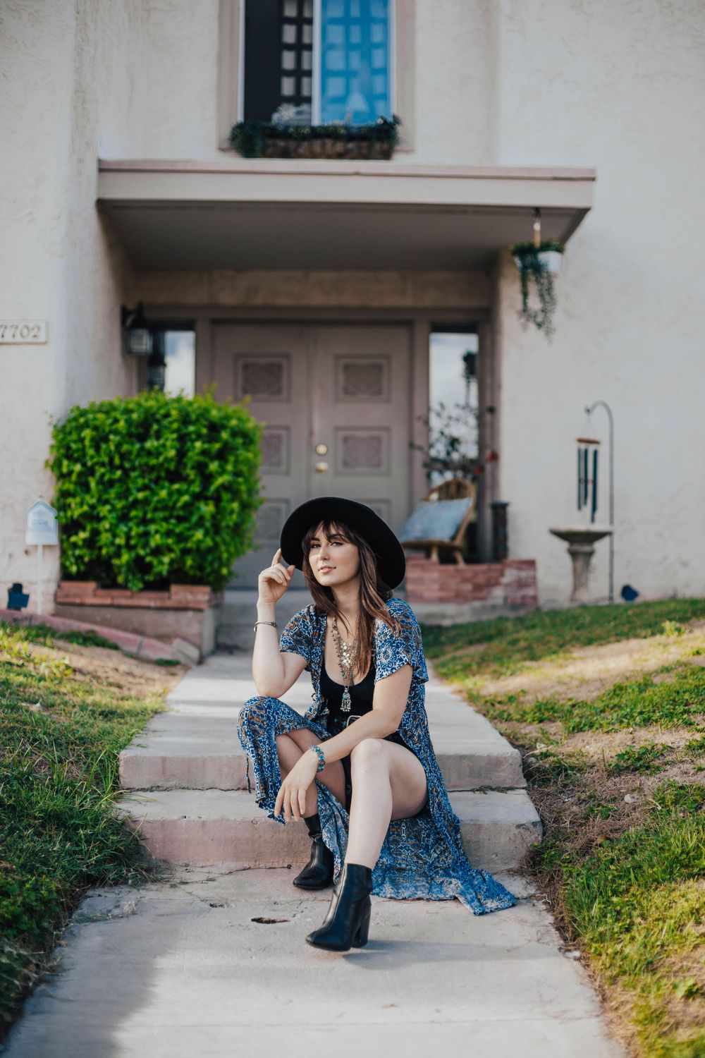 Fashion photoshoot in a blue floral dress and black hat against a white building exterior.