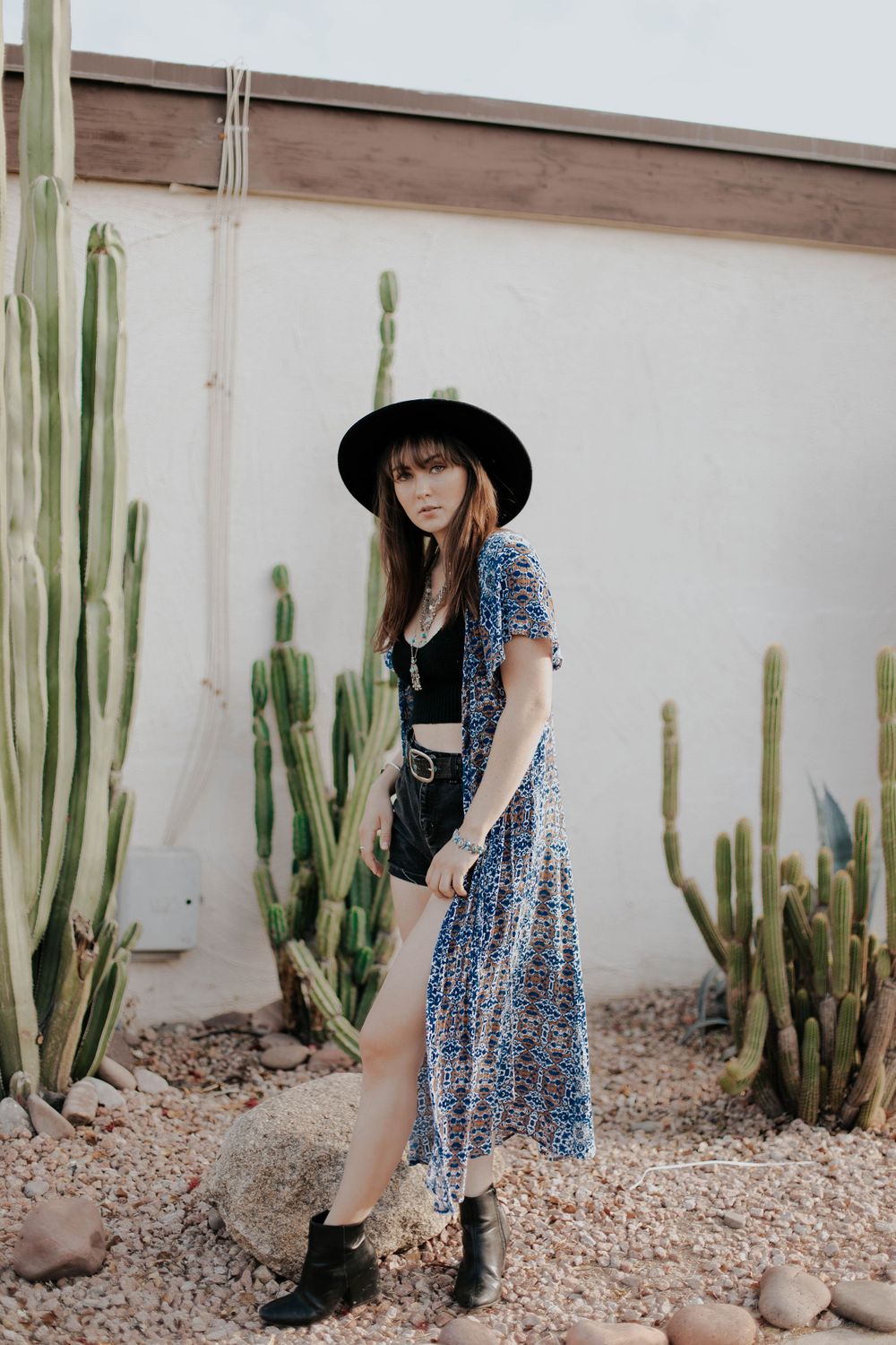 Desert garden photoshoot featuring blue dress and black hat among tall cactus plants.