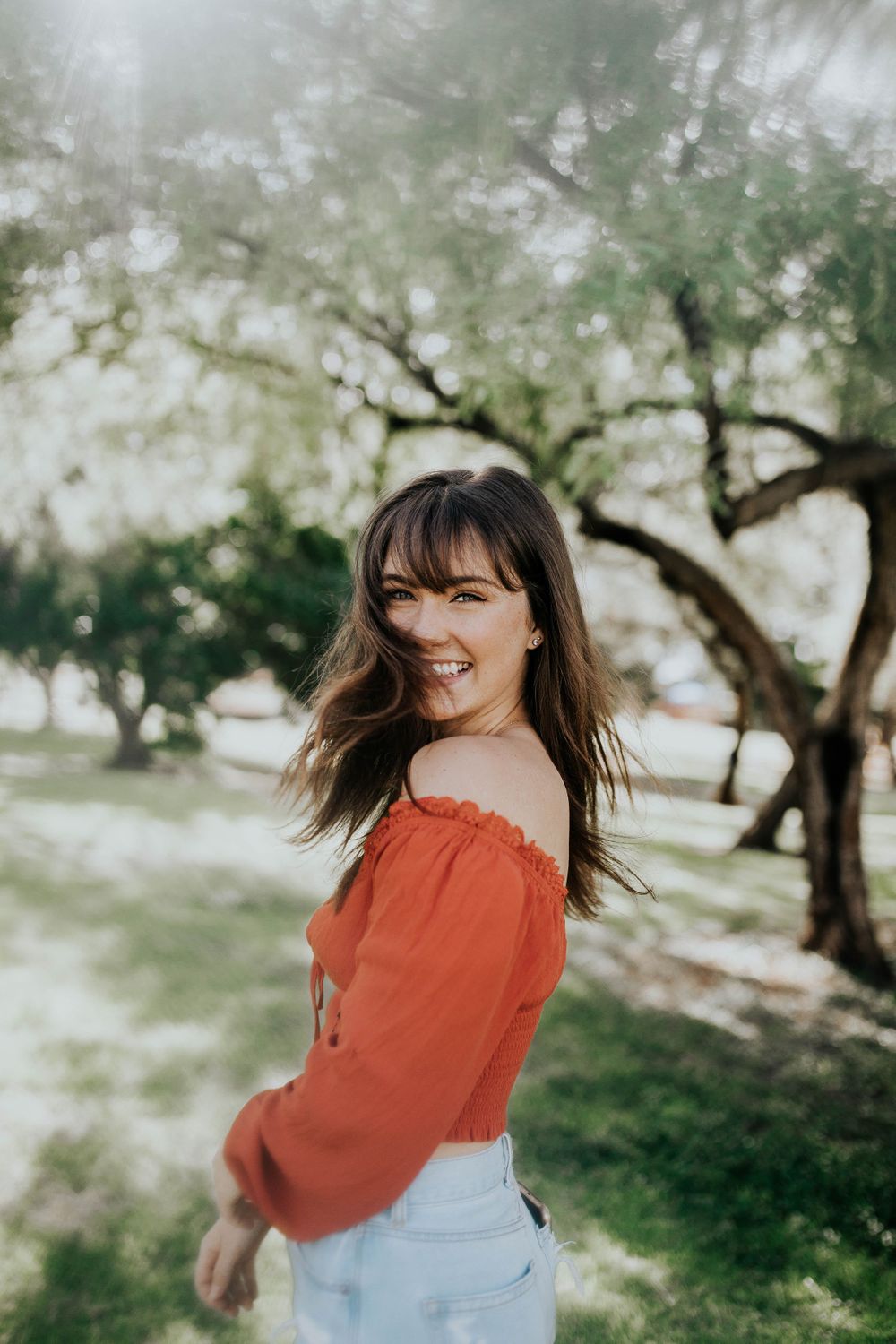 A person in a rust-colored top twirls playfully in a sunlit garden setting with trees in the background.