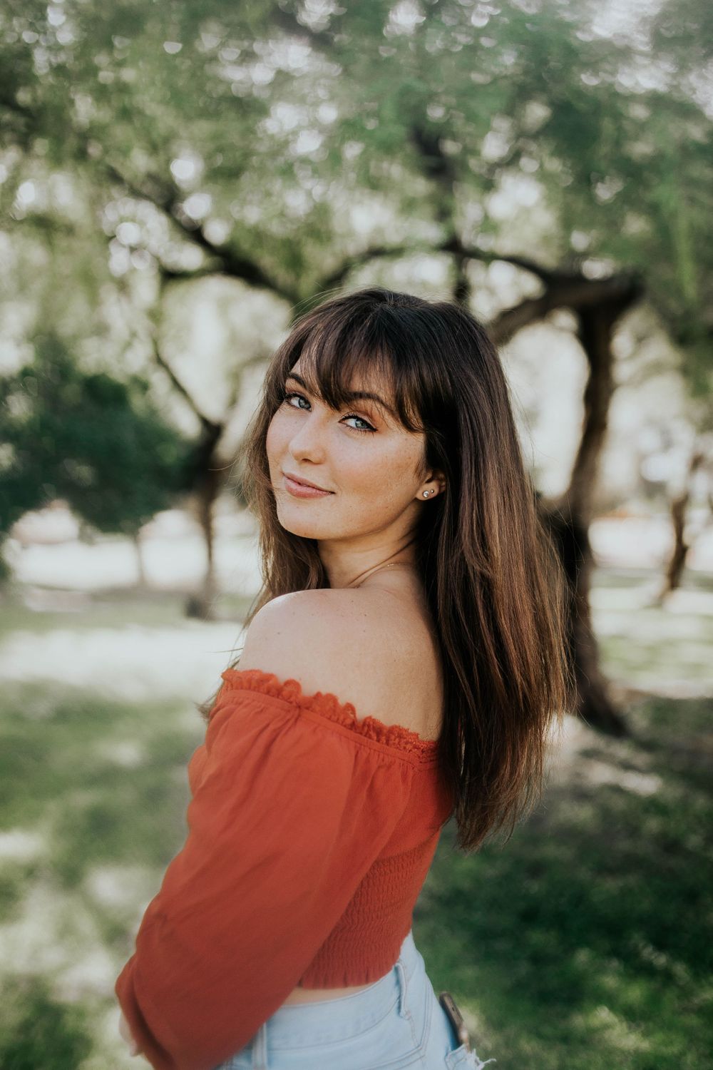 Someone wearing an off-shoulder red top poses casually against a dreamy natural backdrop with trees.