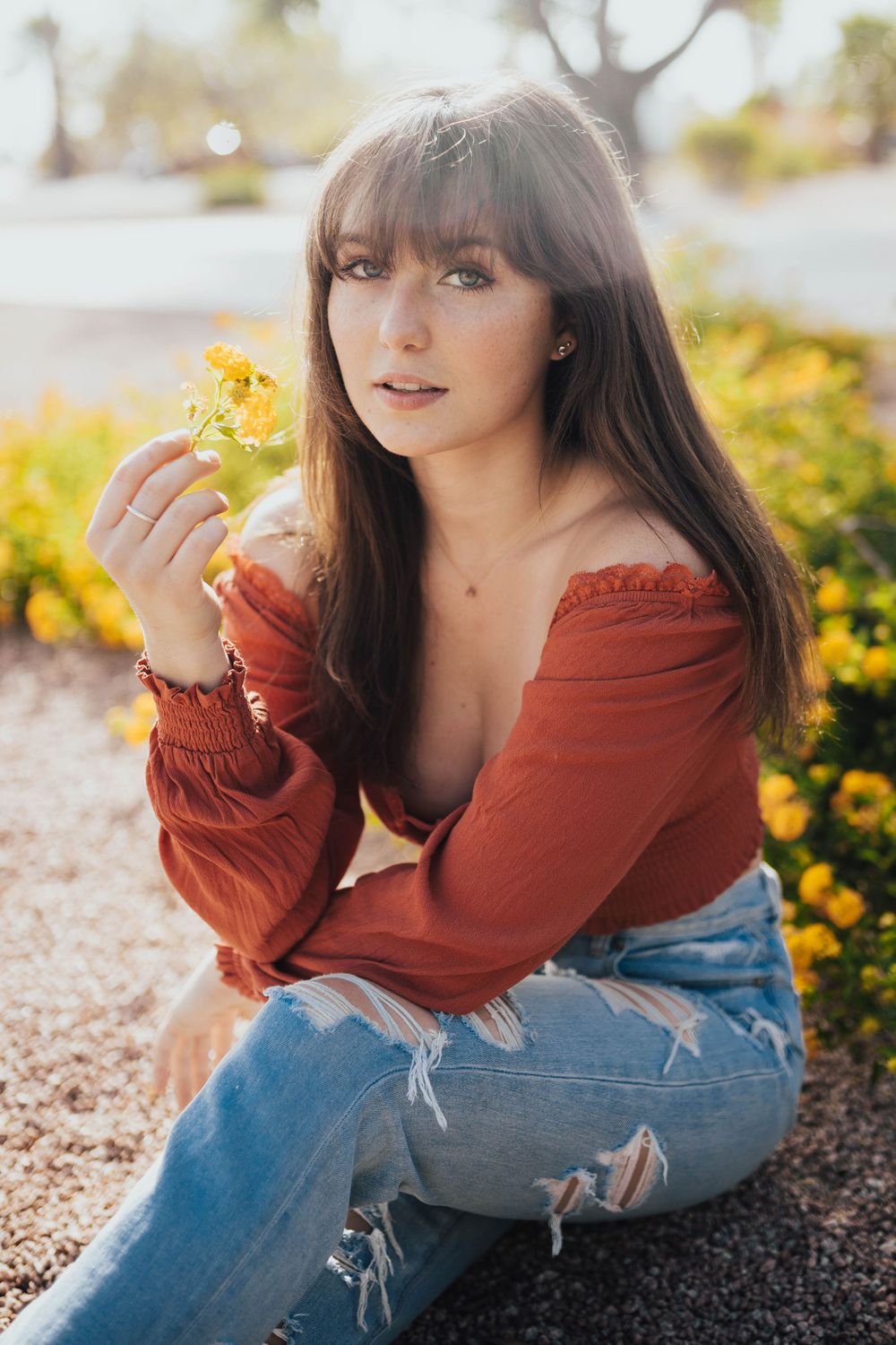 Someone sits among yellow flowers wearing a red off-shoulder top and ripped jeans in warm sunlight.