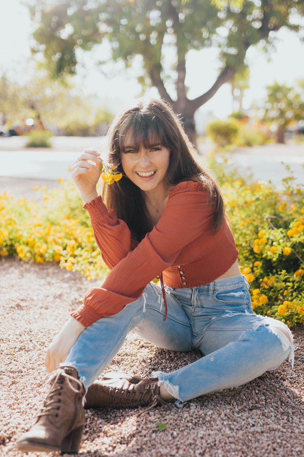 A relaxed outdoor photoshoot on gravel featuring casual denim, brown boots and a rust-colored top.