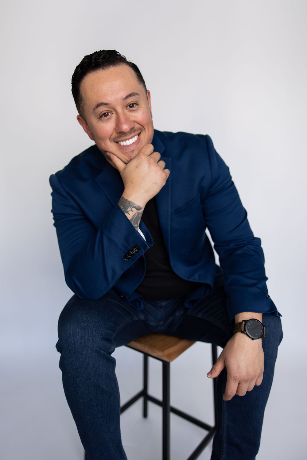 Person in navy blazer and jeans sitting on stool with casual pose and friendly smile.