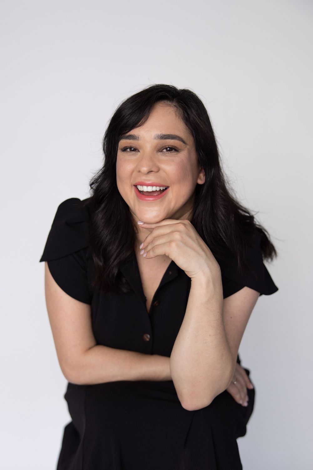 Portrait of person in black dress with dark hair smiling warmly against light backdrop.