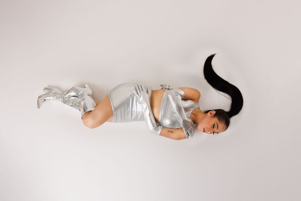 Model in silver outfit poses lying down in minimalist white studio setting.