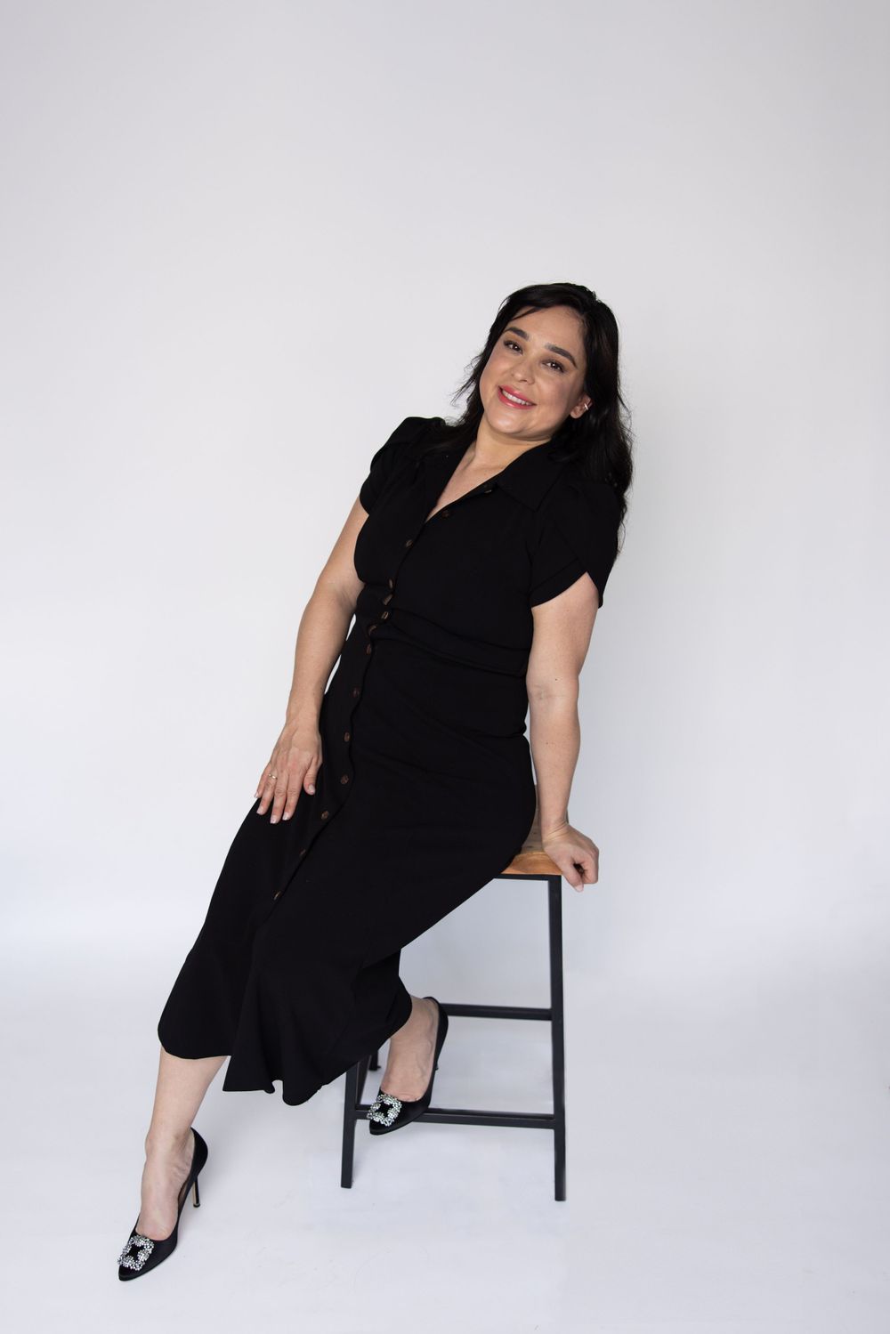 Professional portrait of person in black dress sitting casually on stool against white background.