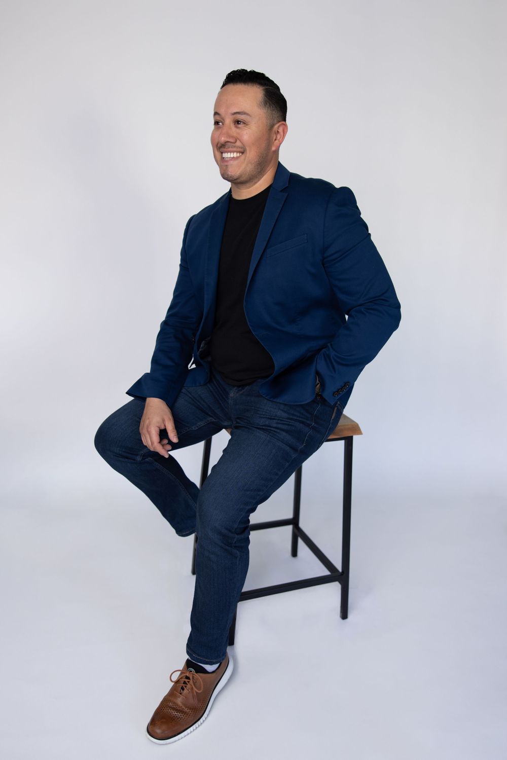 Professional portrait of person in blue suit sitting on stool in studio setting.