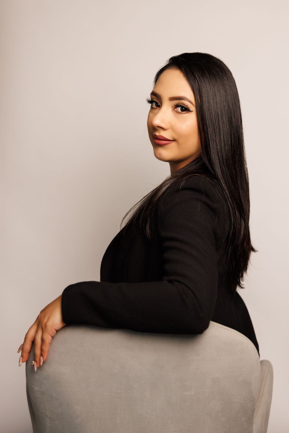 Studio portrait of person in black blazer seated sideways on grey chair.