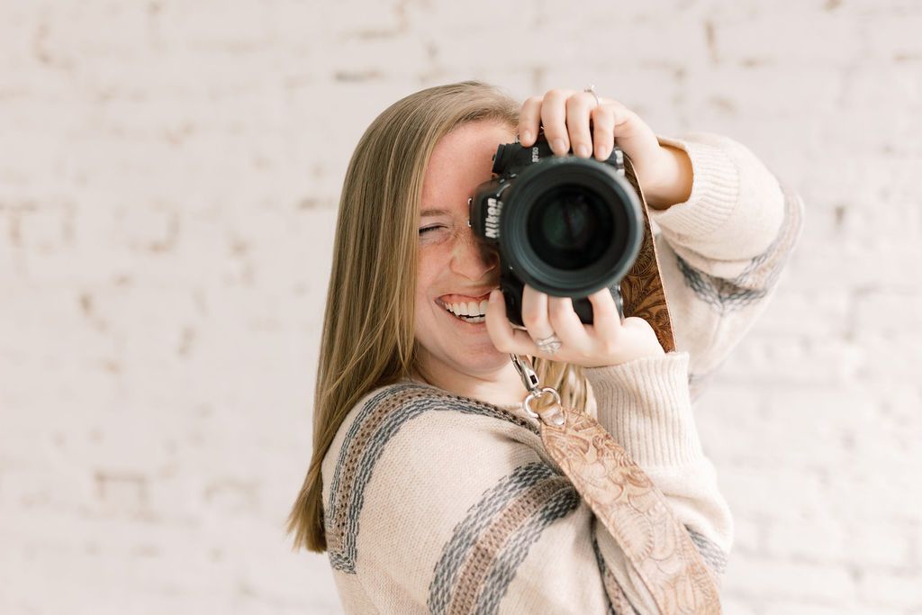 Photographer holding a camera wearing a cream sweater against white brick background.