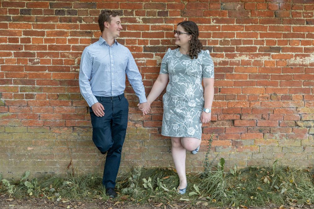 A couple holds hands while standing against a red brick wall in casual attire. Calke Abbey.