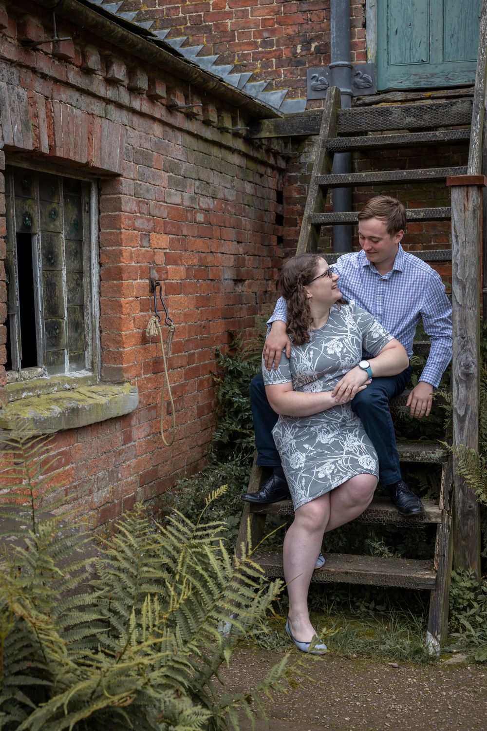 A couple embraces on rustic wooden stairs against a brick wall with ferns growing below.Calke Abbey.