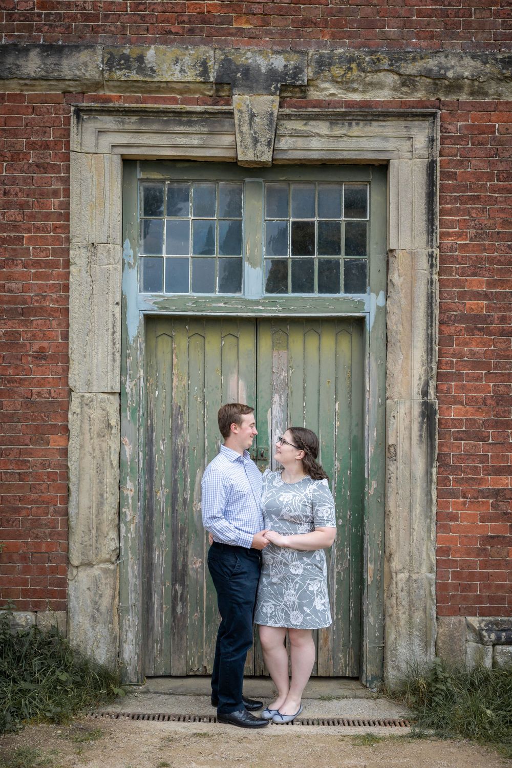 A romantic portrait of a couple standing in front of an old wooden door with brick walls. Calke Abbey.