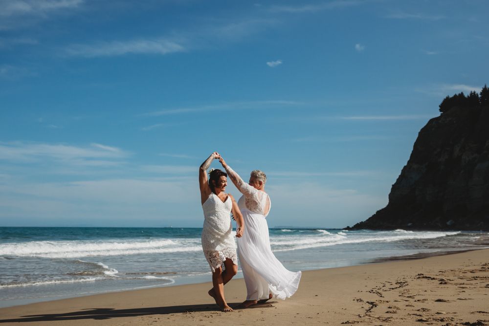brides dancing in white wedding dresses on beach in Napier in sun, same sex wedding photos, by Nelson photographer Lula