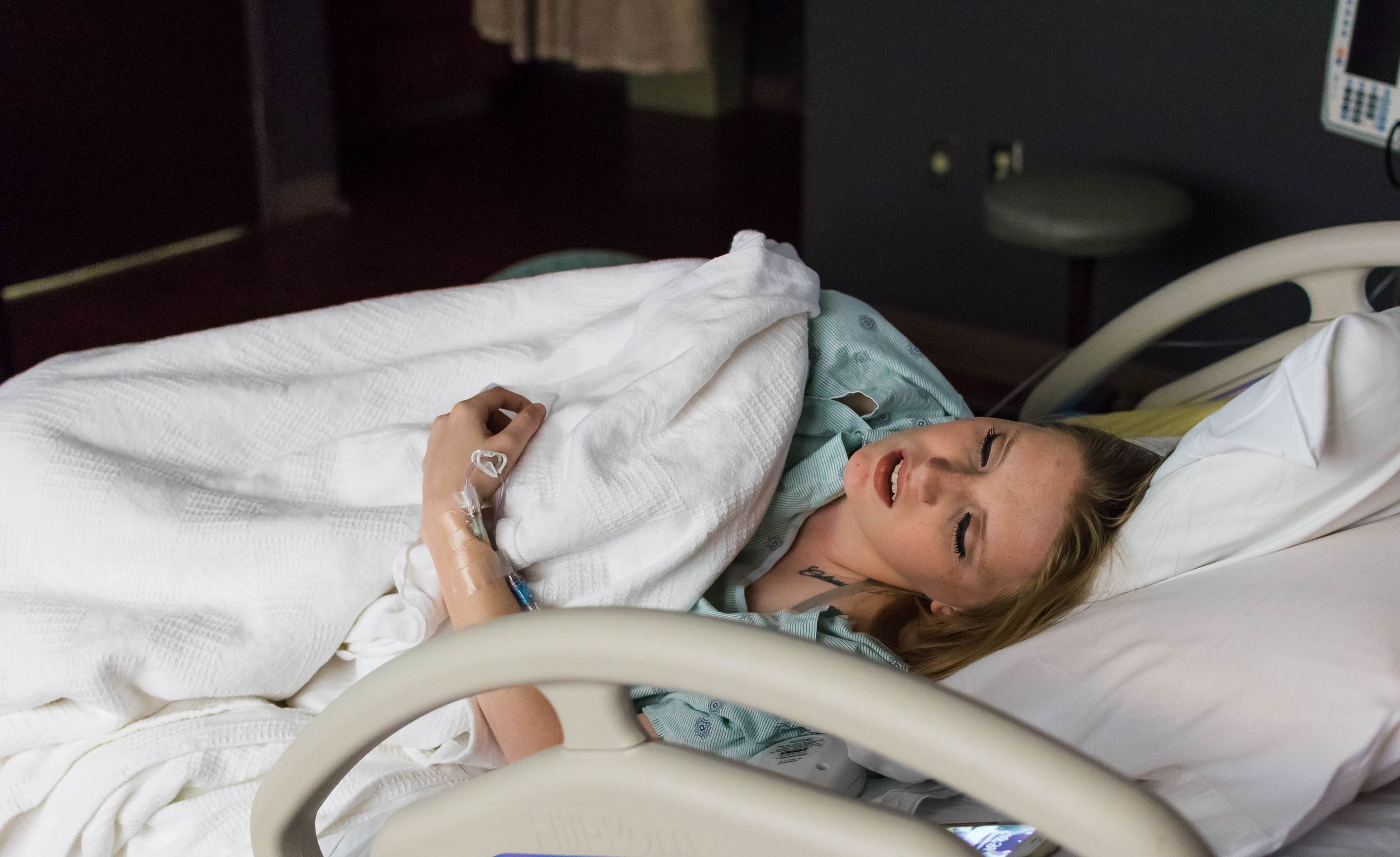 A person in a hospital bed with her eyes closed and moaning as she holds her stomach