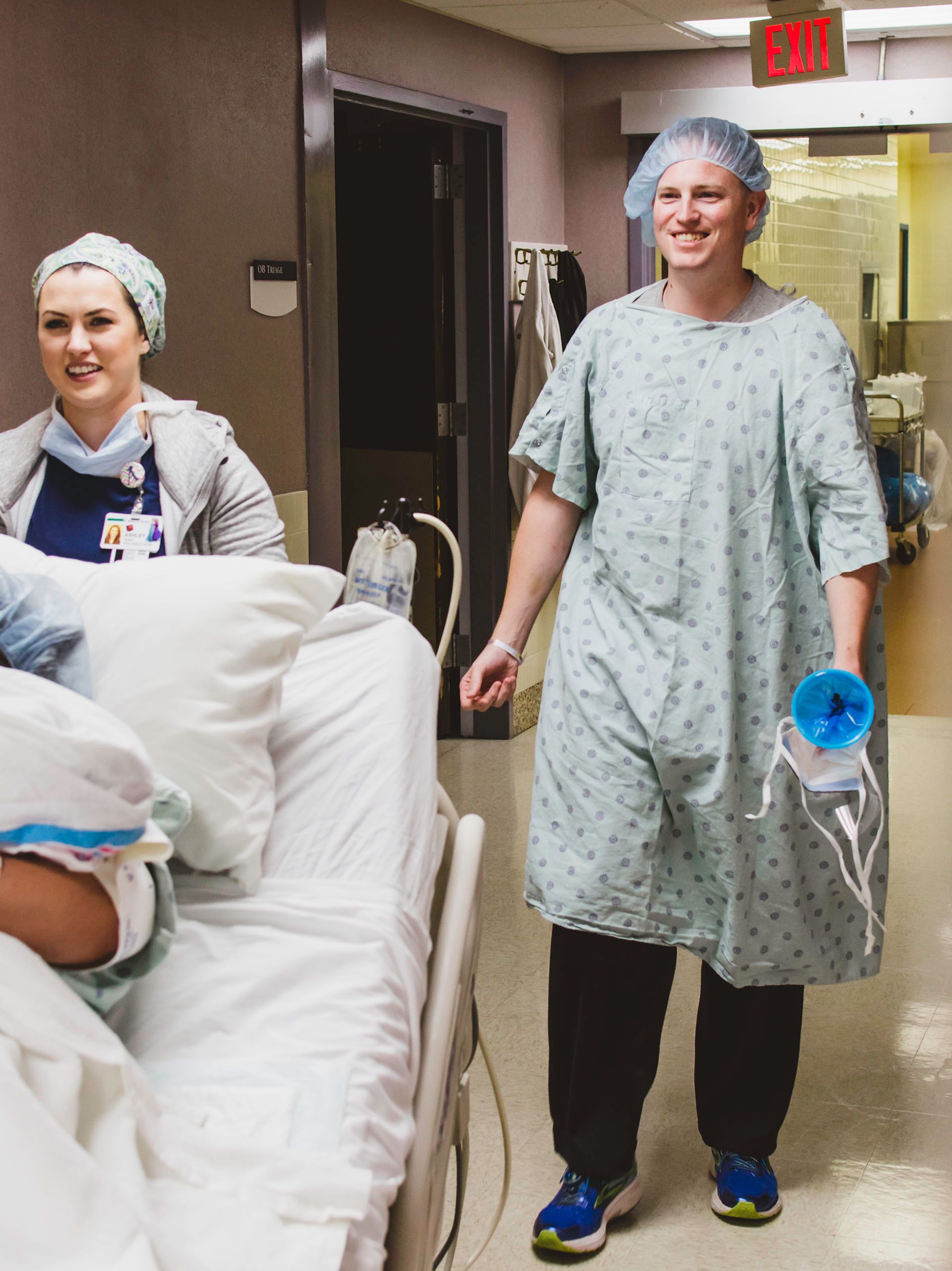 A person smiling while wearing a hospital gown and scrub hat as his wife is wheeled on a hospital bed