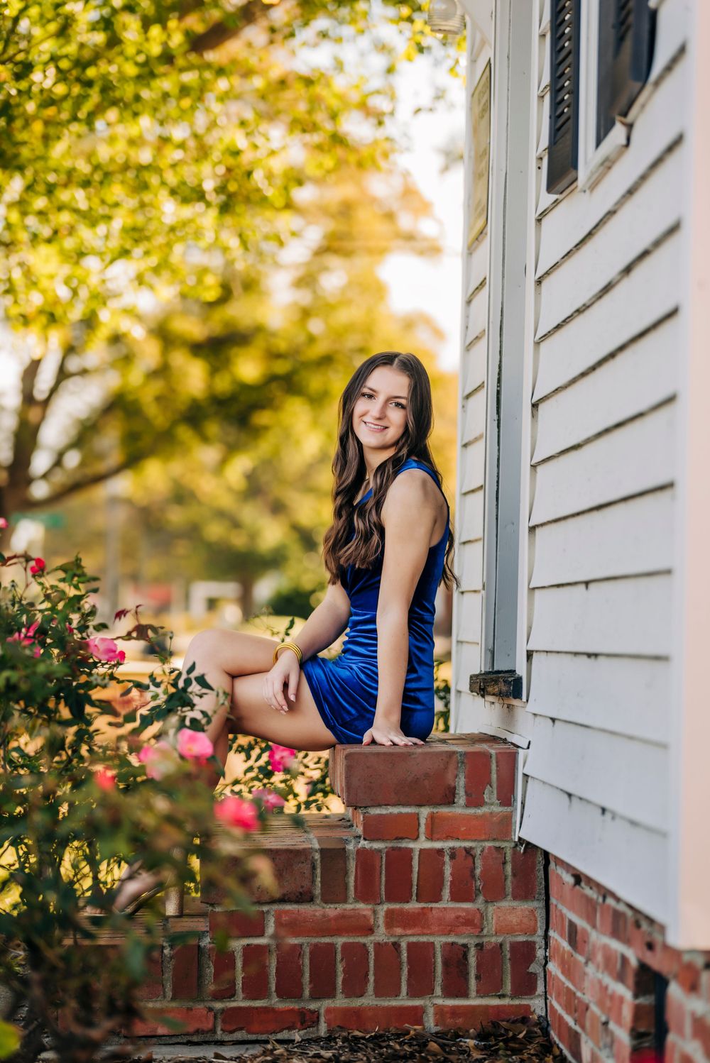 Portrait of young woman in blue dress sitting on brick steps by white house with flowers.