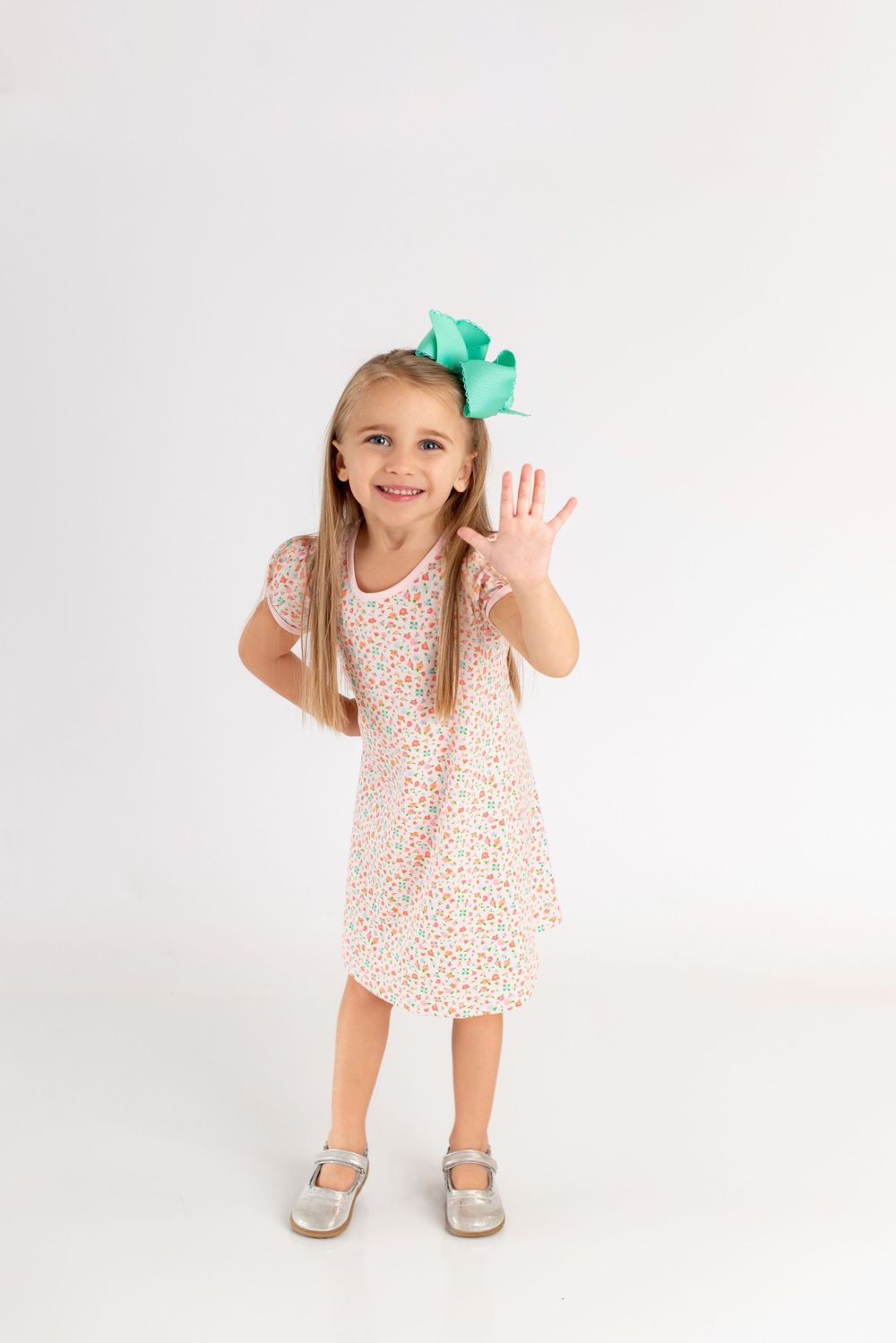 Young girl in floral dress with mint green bow waves at camera against white background.