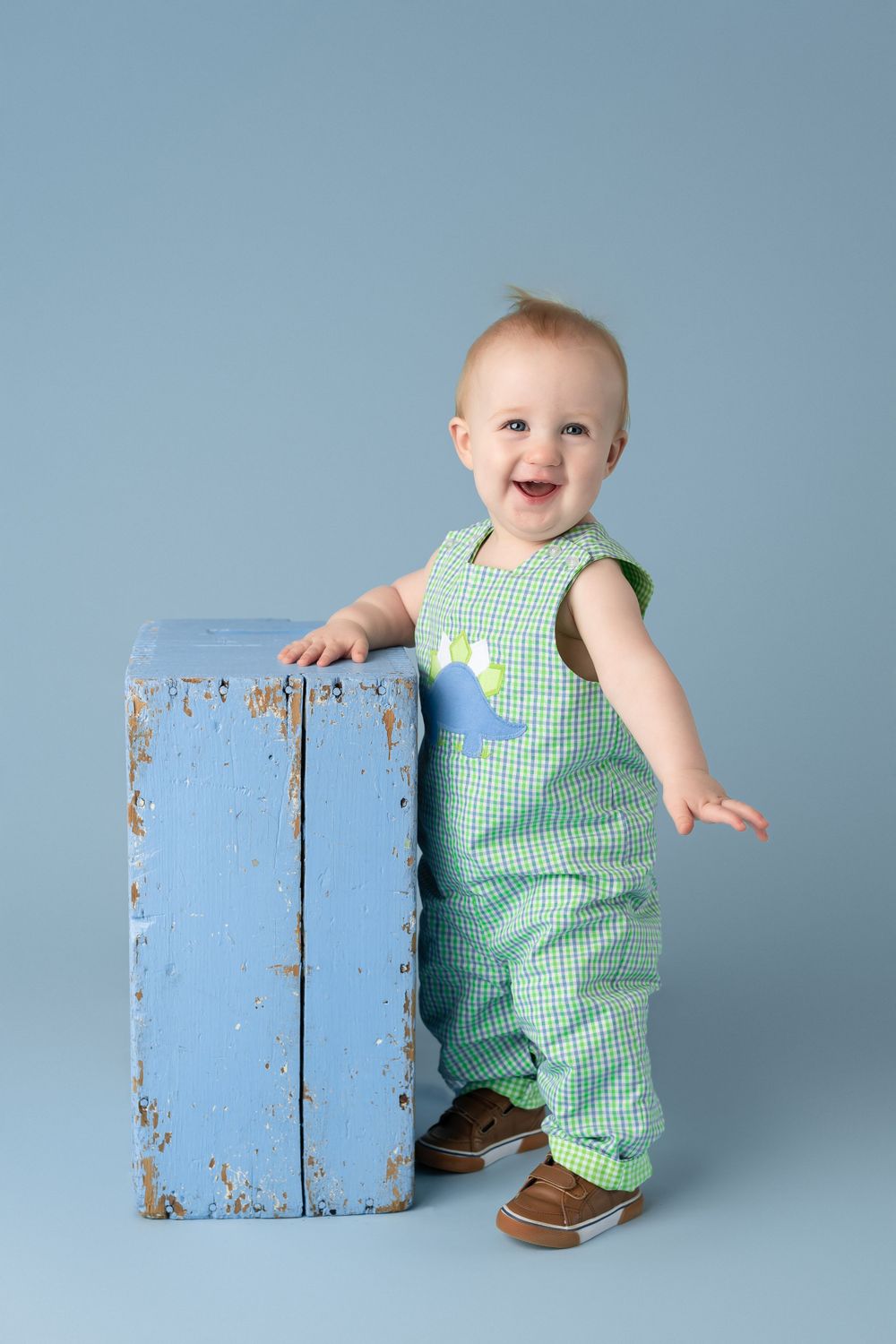 Baby in green gingham overalls stands next to distressed blue wooden box against blue backdrop.