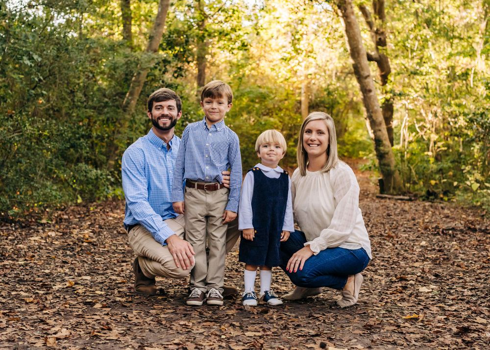 Portrait of a young family of four sitting together on fallen leaves in a wooded setting.