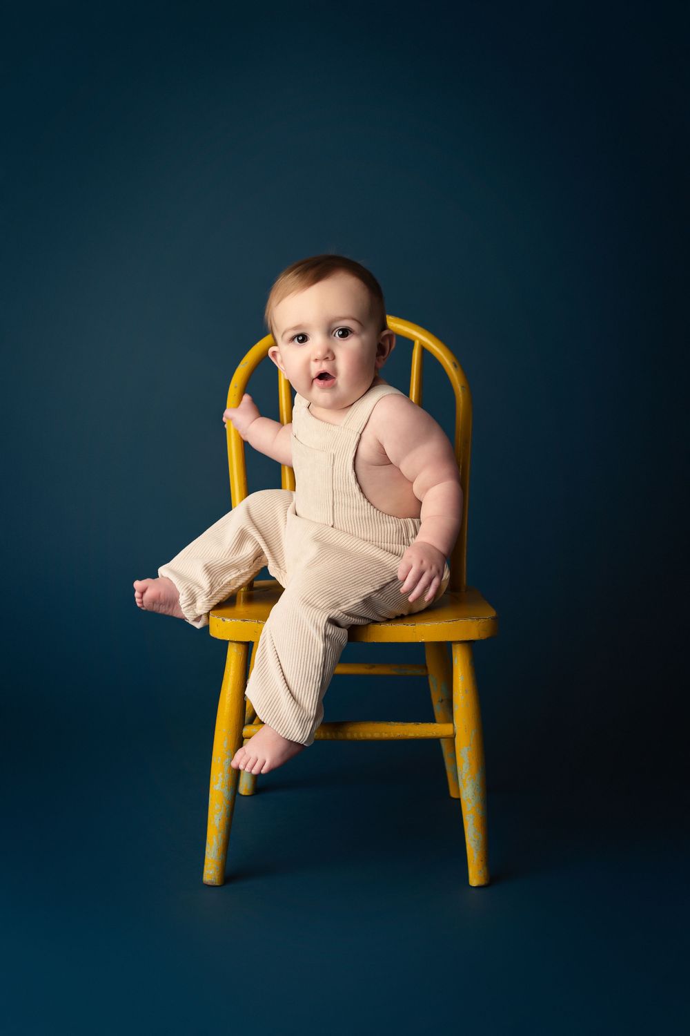 Baby in cream-colored outfit sits on yellow wooden chair against deep blue background.