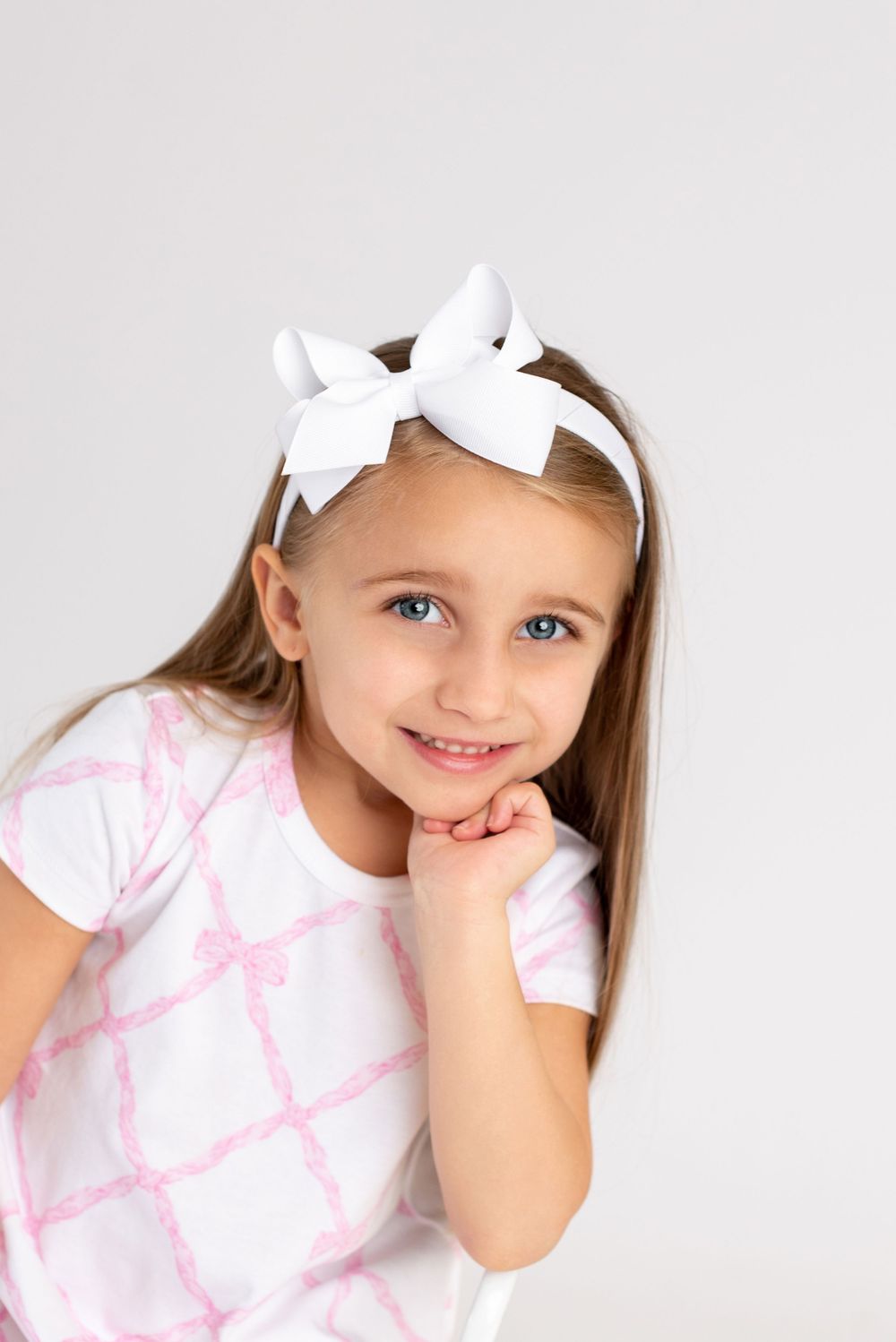 Young girl wearing white bow headband and pink patterned shirt poses against white background.