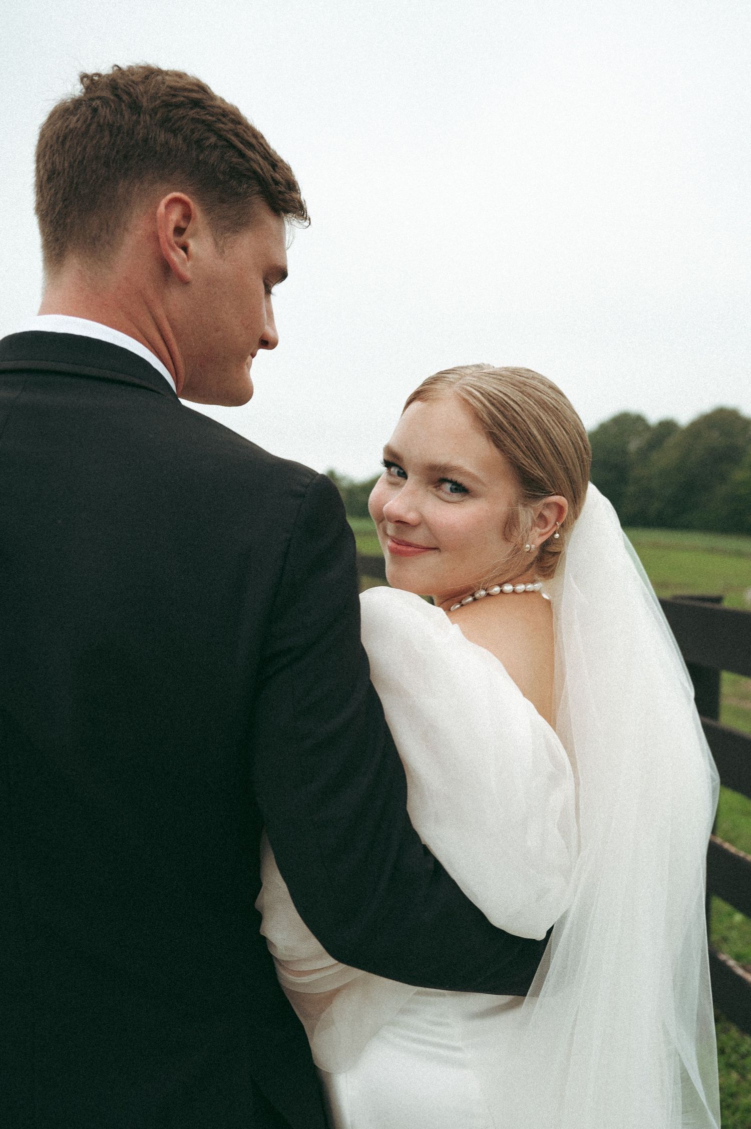 Bride in white dress and veil looks back while embraced by groom in black suit.