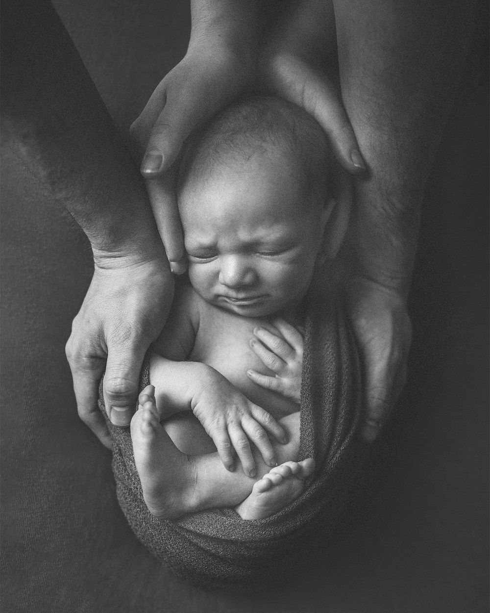 Black and white newborn portrait of a crying baby cradled gently in parents loving hands.
