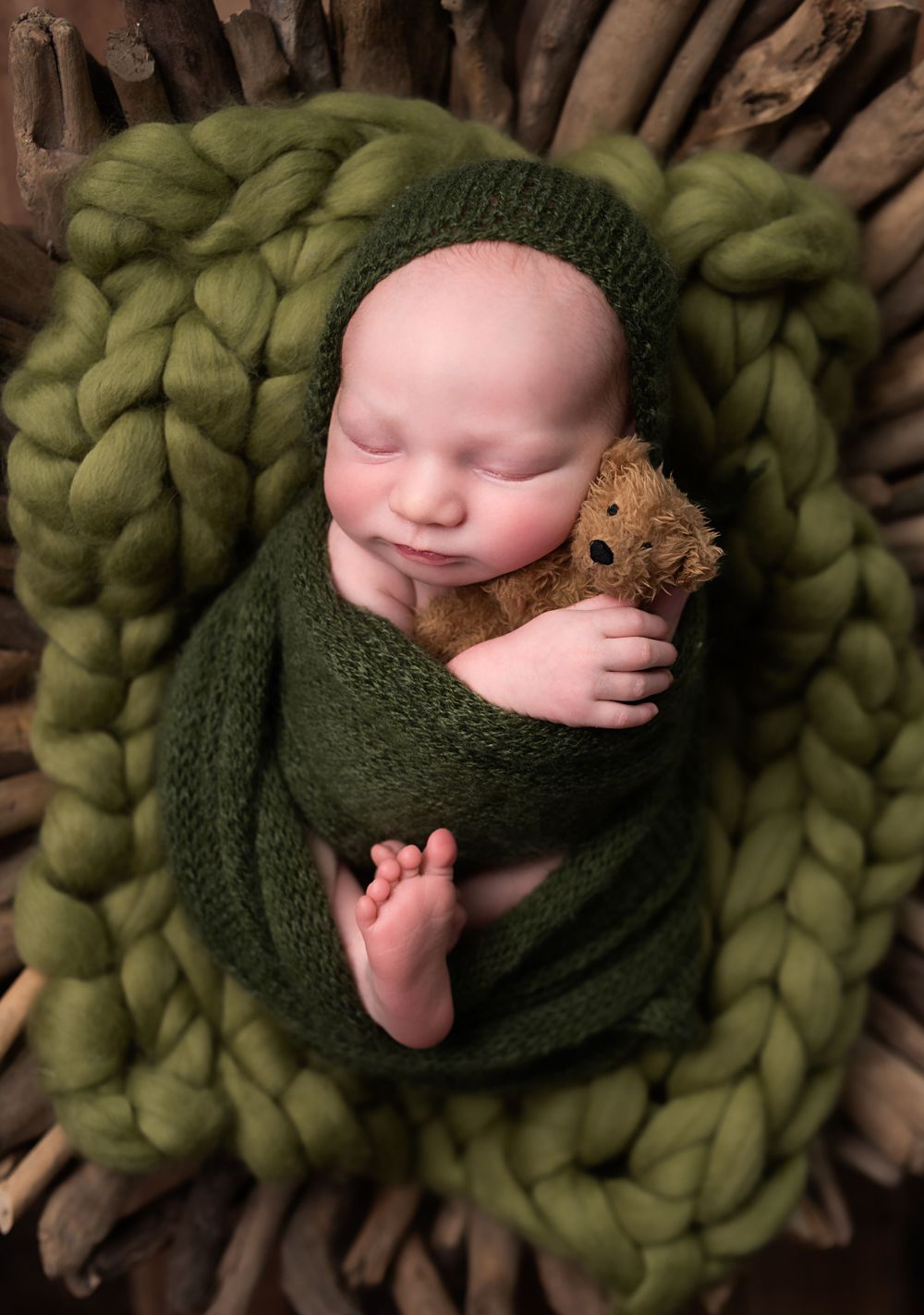 Sleeping newborn baby wrapped in green knit blanket holding teddy bear in rustic wooden basket.