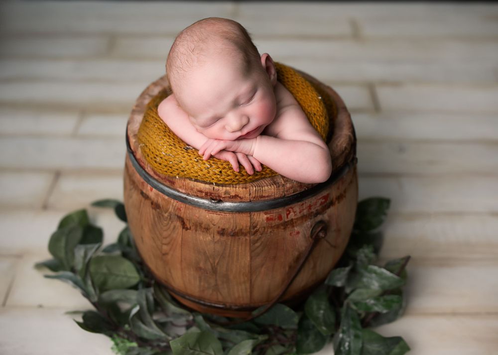 Adorable sleeping newborn baby posed in a rustic wooden barrel with yellow knit and green leaves.