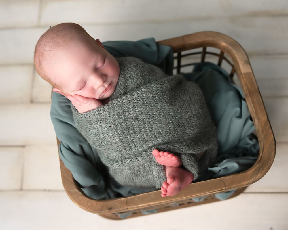 Sleeping newborn baby wrapped in green knit swaddle with tiny toes peeking out of wooden basket.