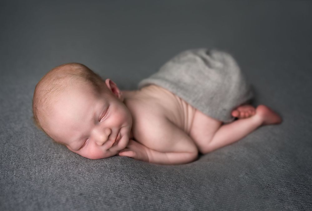 Peaceful newborn baby sleeping on stomach with hands under cheek on a soft gray blanket.