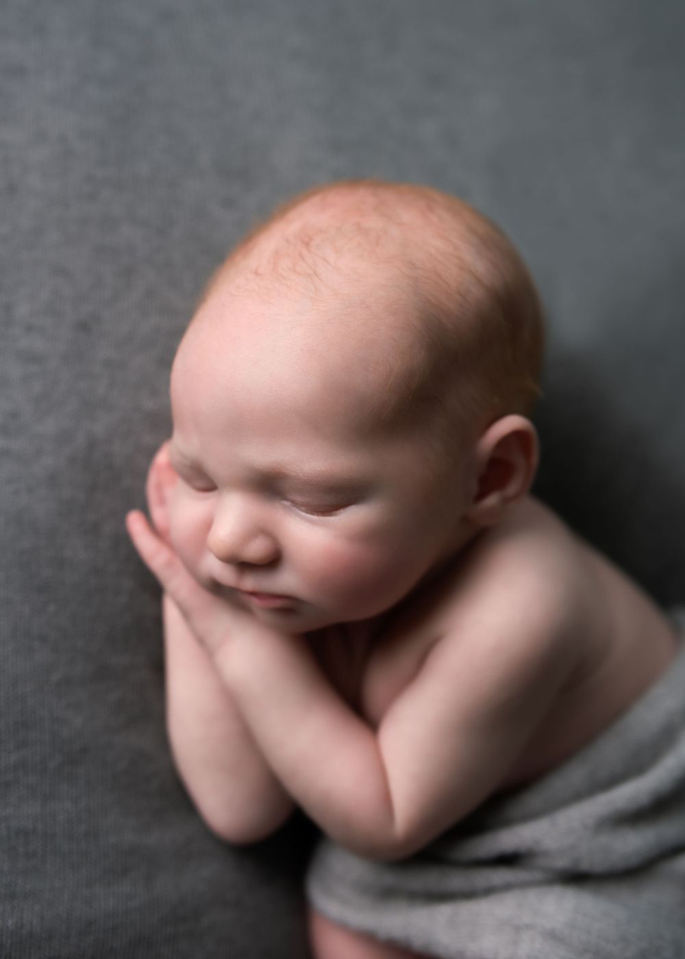 Close-up portrait of a sleeping newborn baby resting on folded hands on a gray backdrop.