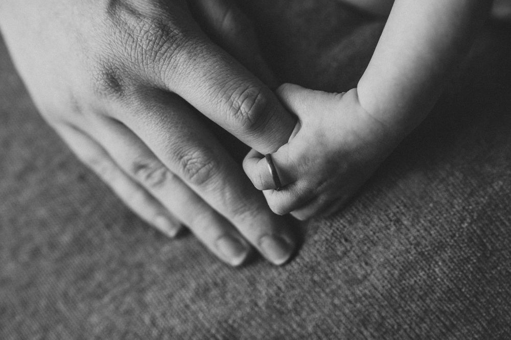 Black and white photo of a newborn babys tiny hand grasping a parents fingers tenderly.