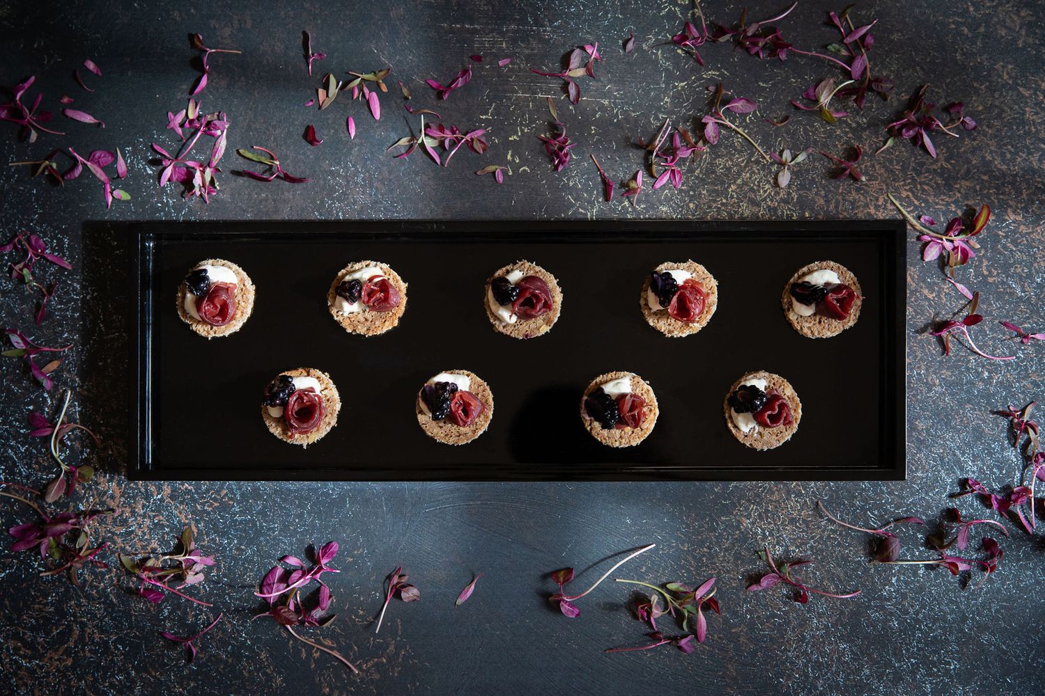 Black platter of elegant dessert bites garnished with purple flower petals from a product photoshoot.