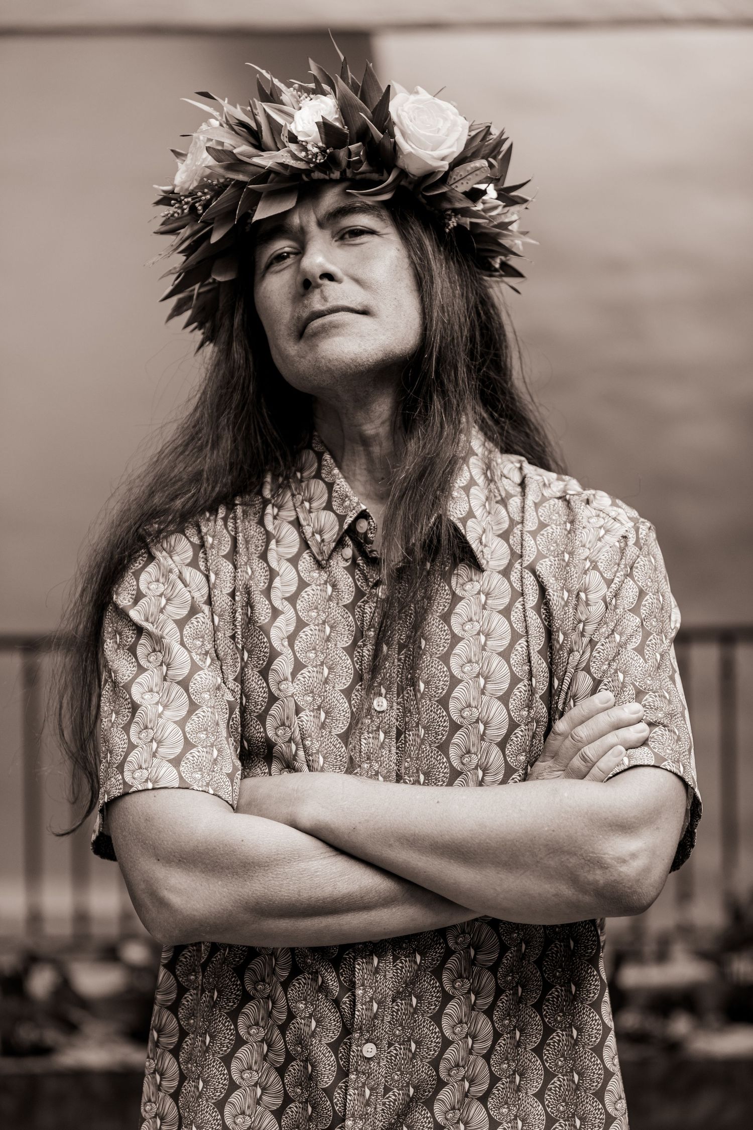 Sepia portrait of person wearing floral lei crown and patterned shirt with arms crossed.
