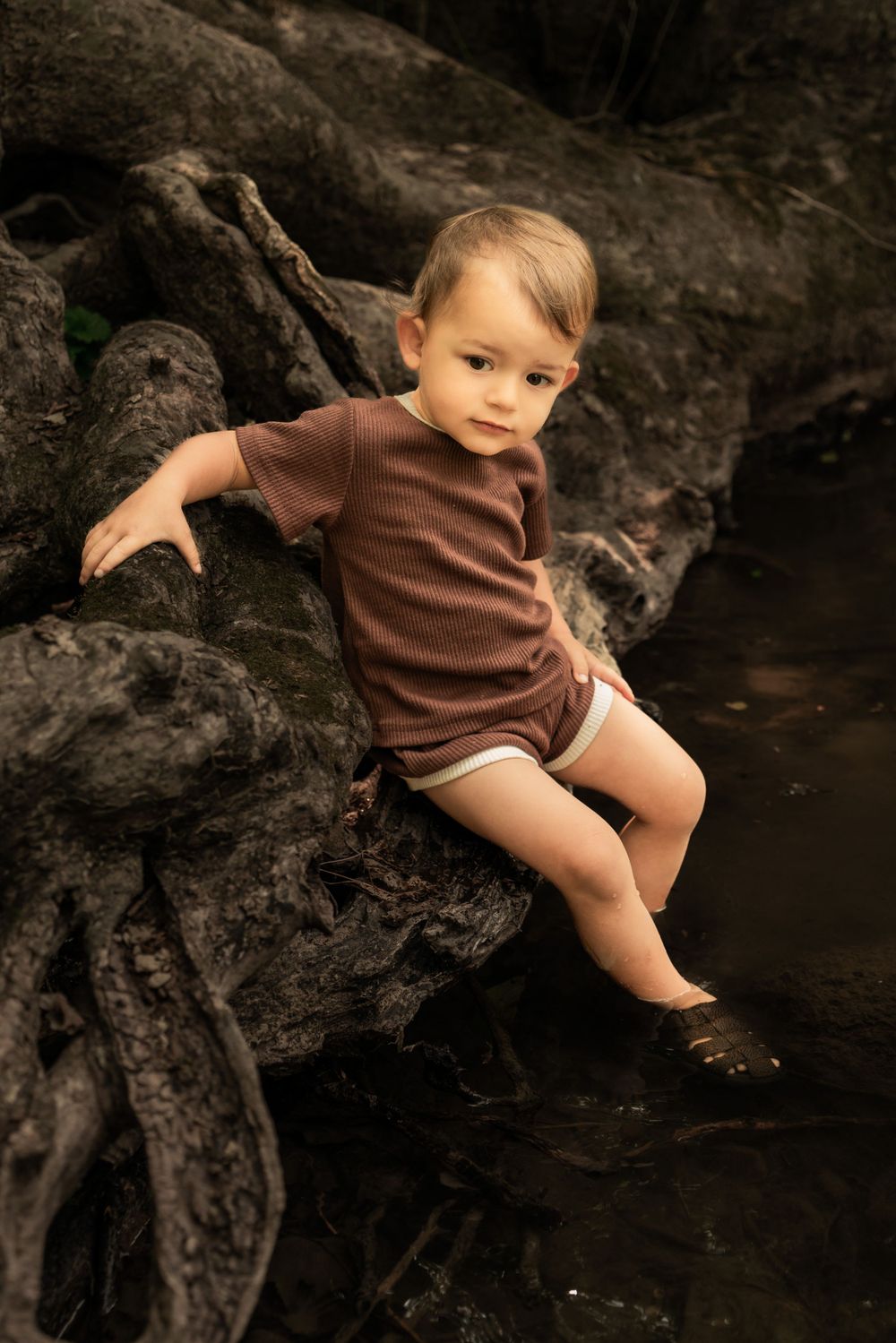 toddler sits on tree roots with feet in river