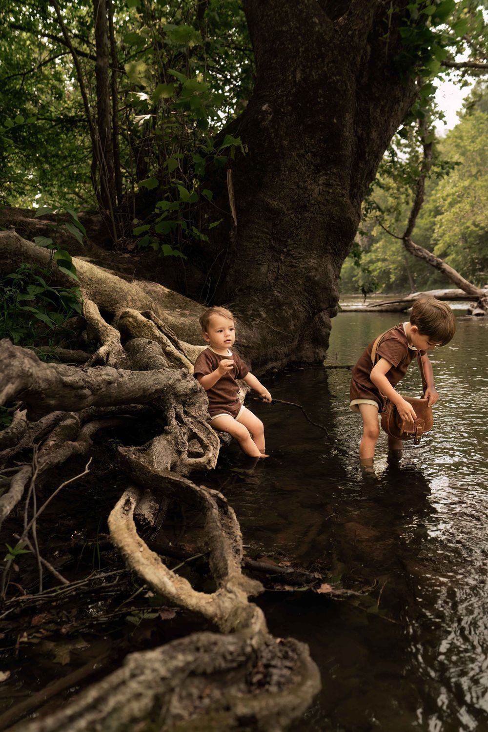 little toddler sits on log by river while big brother looks for rocks