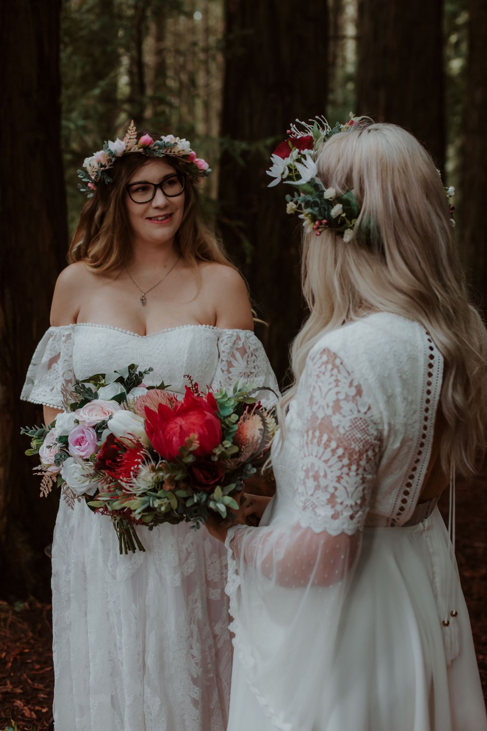 Two people wearing white lace dresses hold floral bouquets in a forest setting.