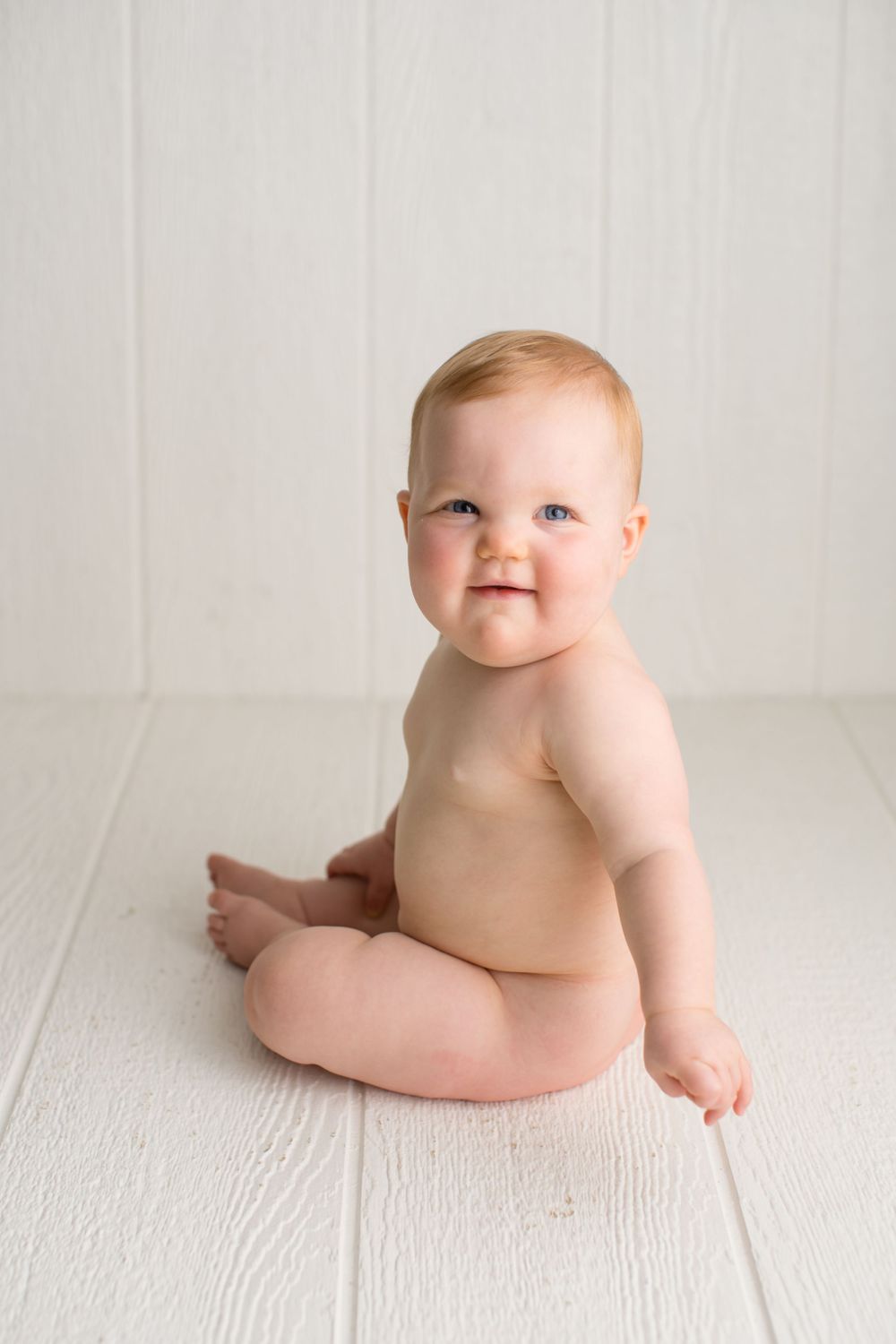 A cheerful baby sitting on white wooden floor during portrait session.