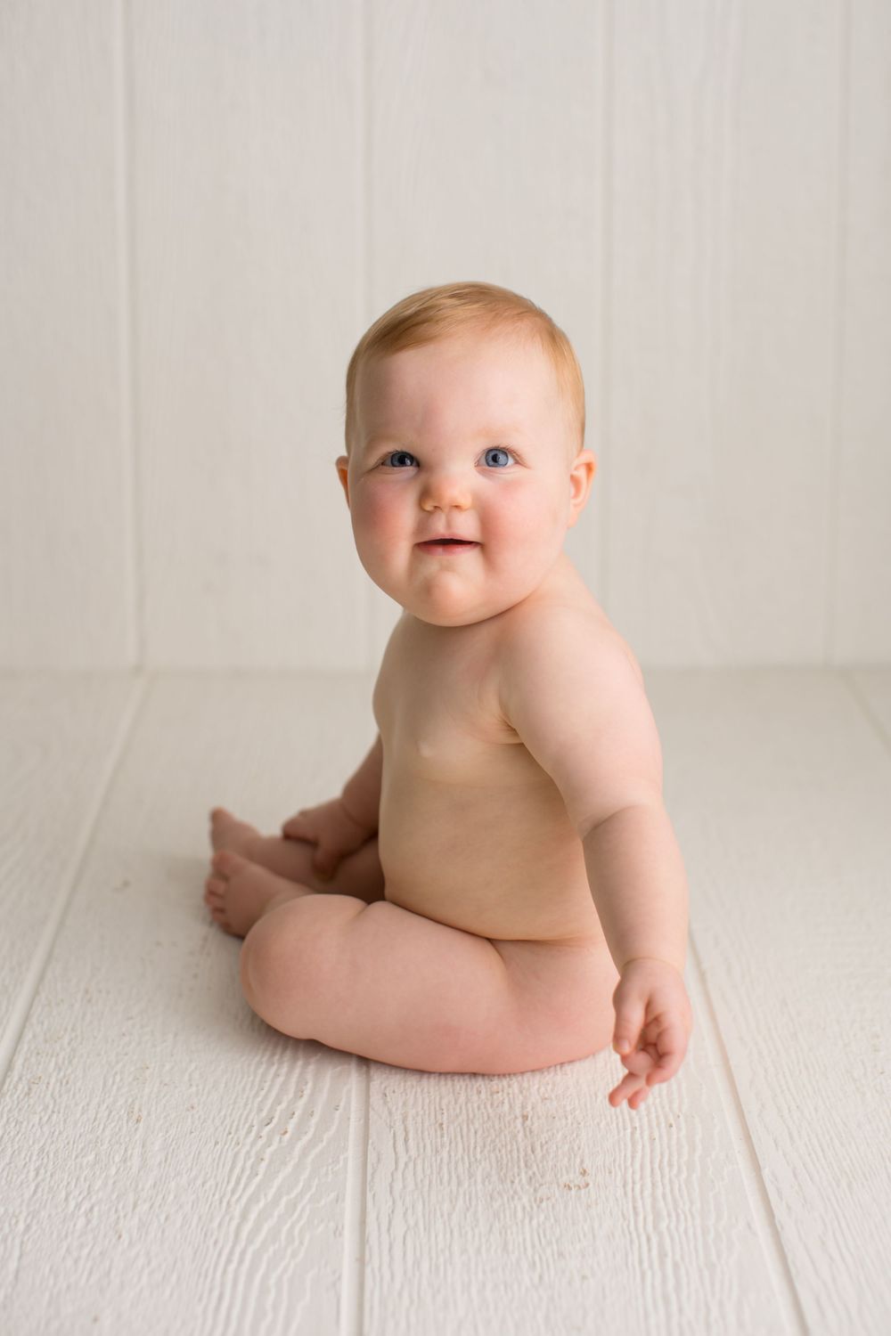 A joyful baby sitting upright on white wooden flooring during studio photography.