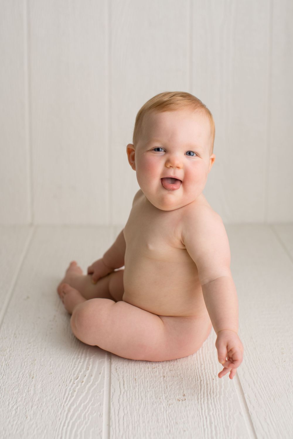 A laughing baby sitting on white wooden flooring during minimalist photo shoot.