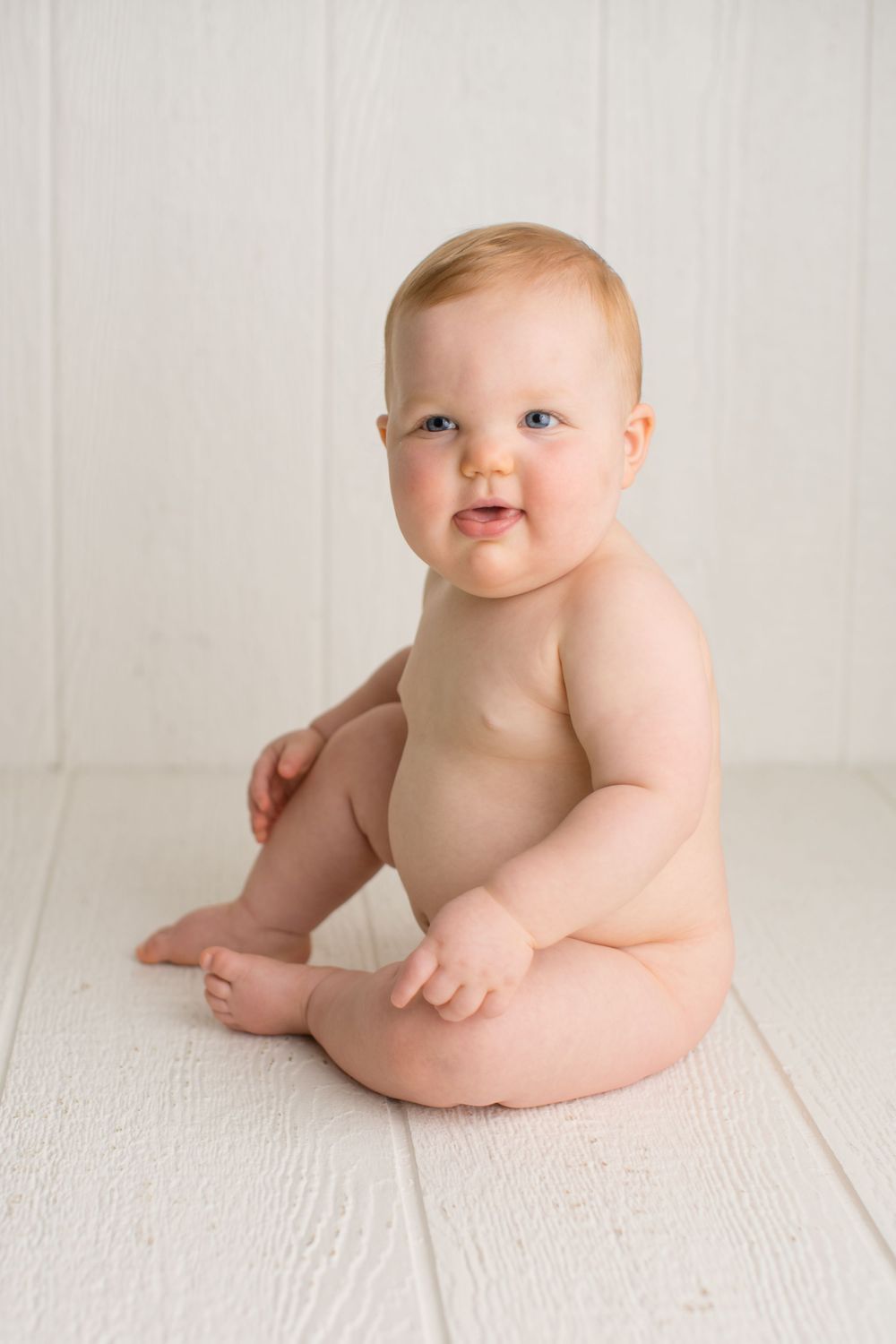 Smiling baby wearing only a diaper sits on white wooden floor for professional portrait.