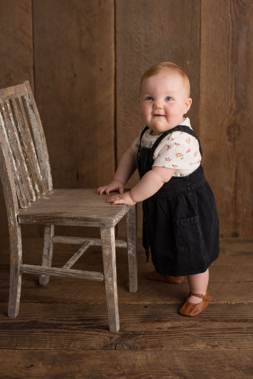 Baby in black overalls and white shirt stands holding vintage white chair against rustic backdrop.