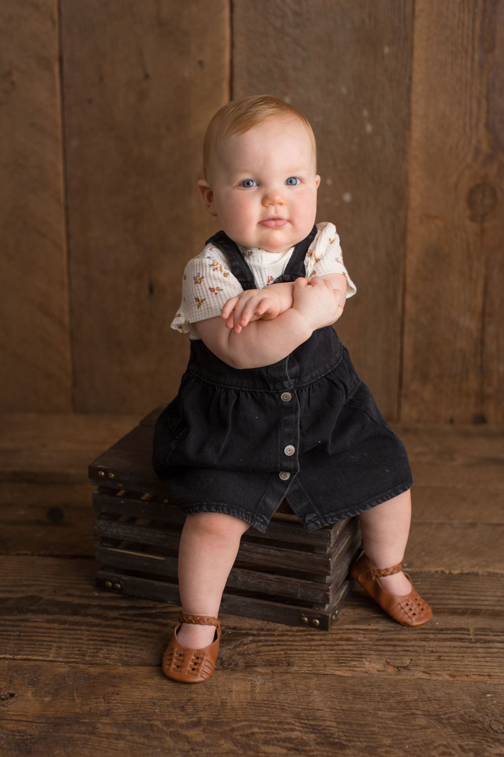 Baby wearing black dress and brown shoes sits on wooden crate in rustic setting.