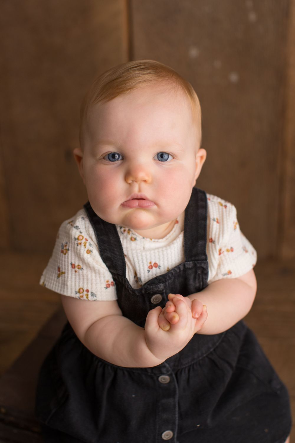 Baby in black dress sits on wooden box against rustic brown wall for portrait.