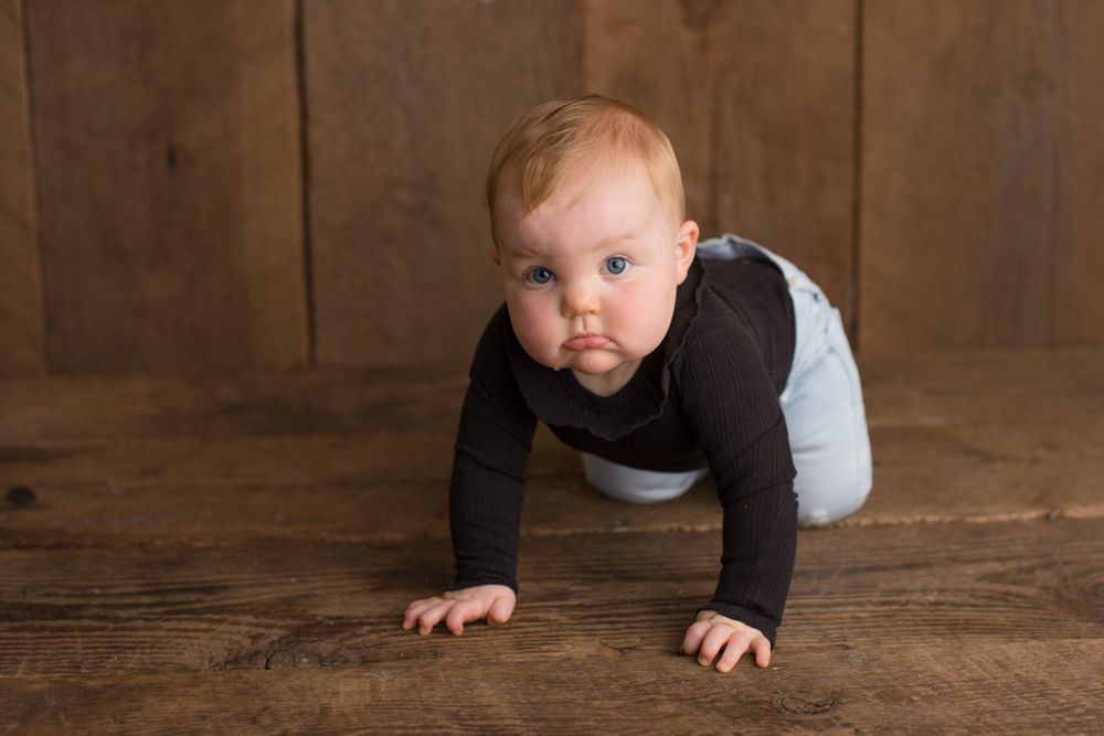 Baby in black top and light pants crawls on wooden floor during photo session.